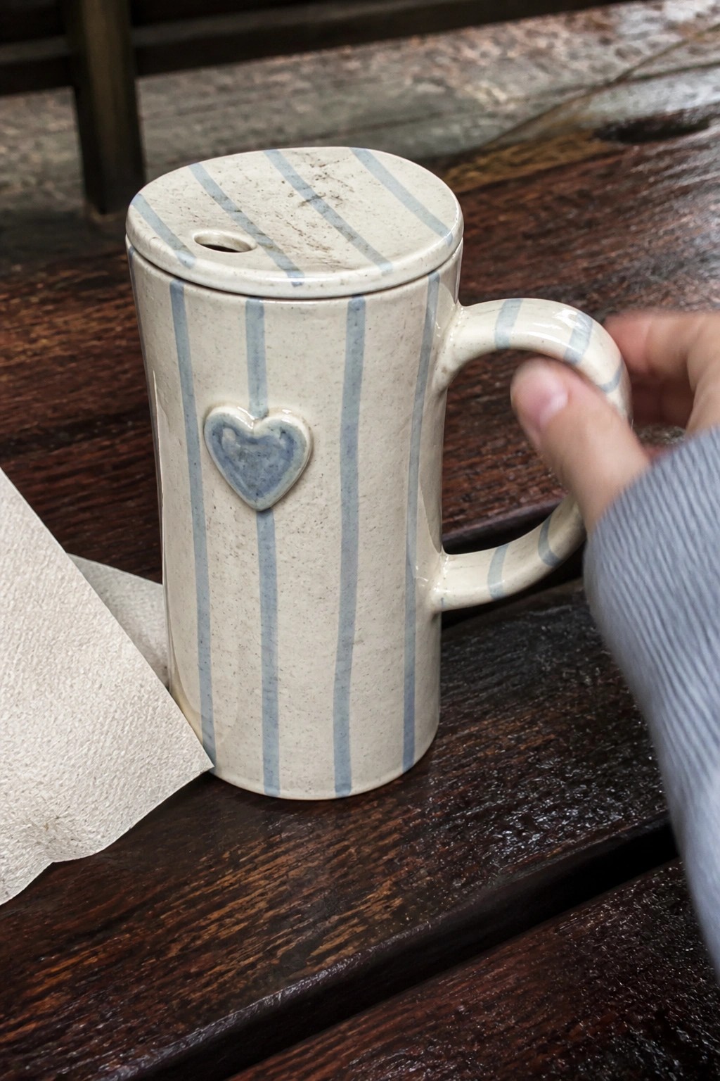 Hand in striped sleeve holding lidded blue-striped ceramic mug with heart on wood table