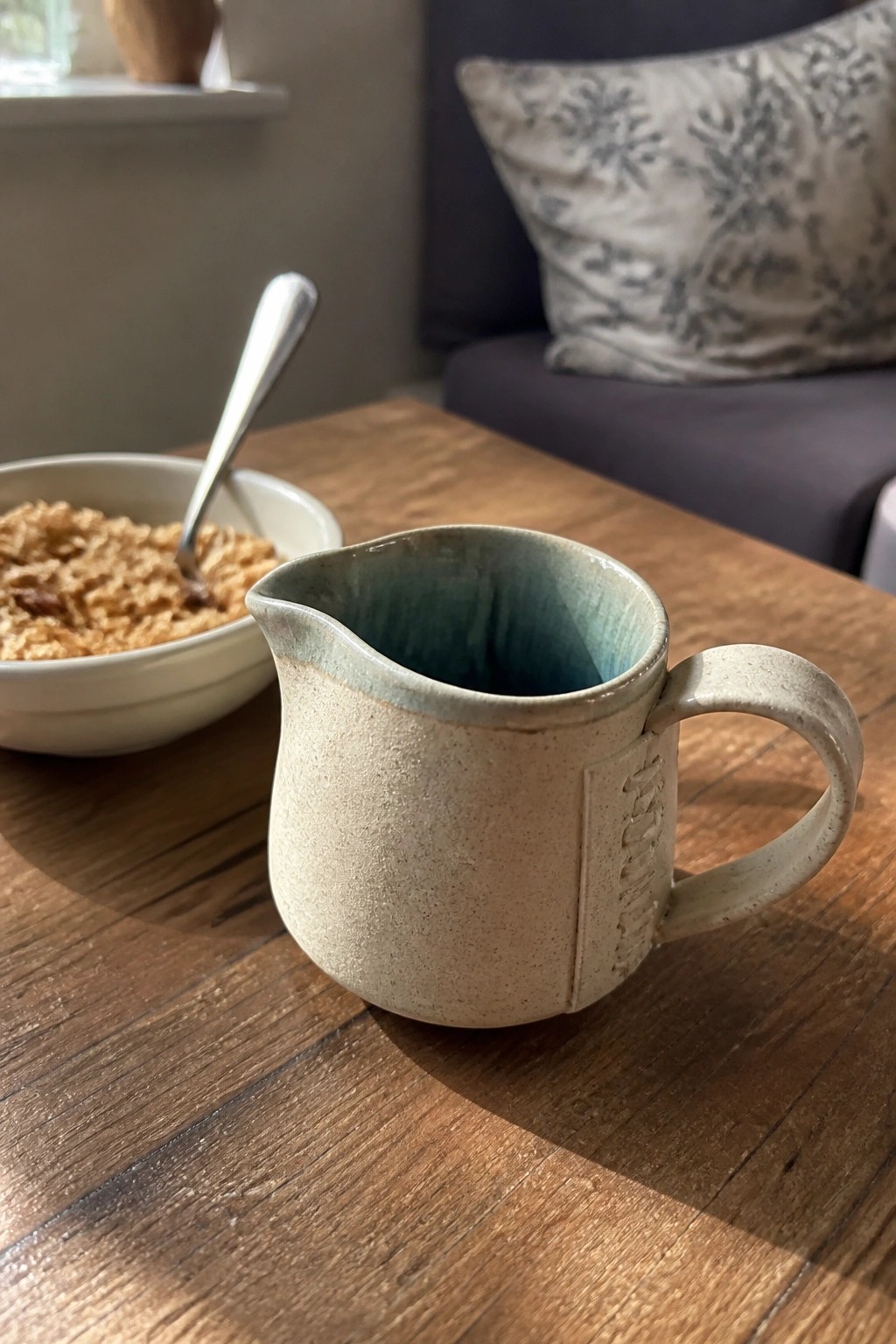 Handmade ceramic pitcher with blue-green interior beside oatmeal bowl on wooden table