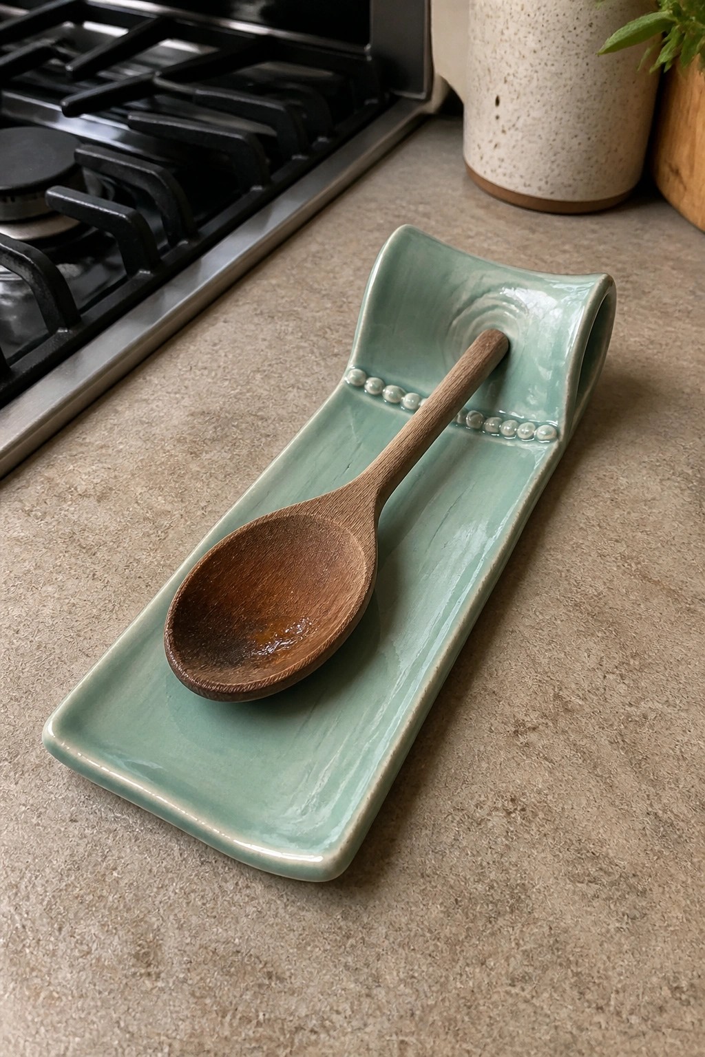 Wooden spoon on pale green ceramic rest with beaded edge on kitchen counter.