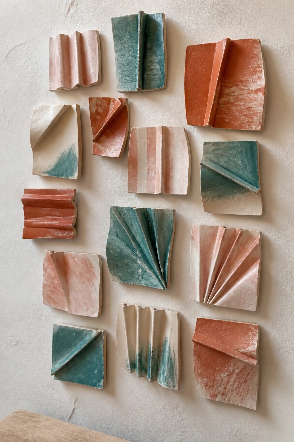 Assorted folded ceramic tiles in pink, teal, and orange hues mounted on a wall.