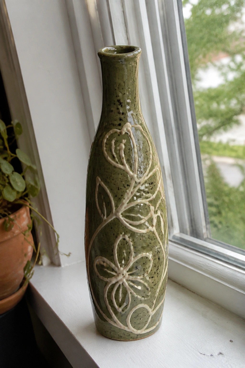 Green ceramic vase with white floral patterns on white windowsill beside plant