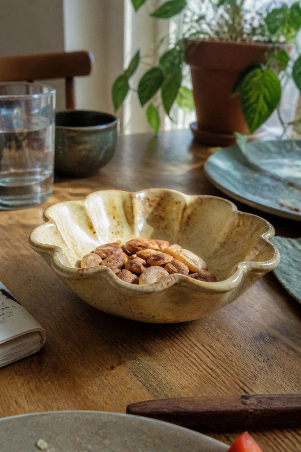 Scalloped beige ceramic bowl of almonds on wooden table with potted plants nearby