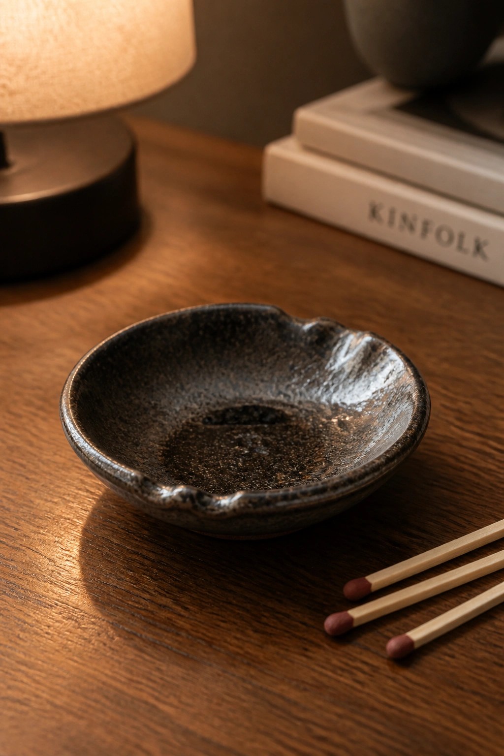 Dark speckled ceramic bowl on wooden table beside matches, lamp, and books