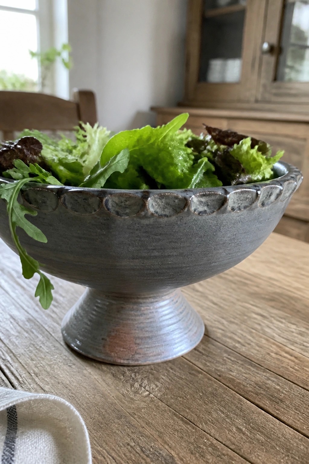 Gray ceramic pedestal bowl brimming with fresh mixed salad greens on wood table