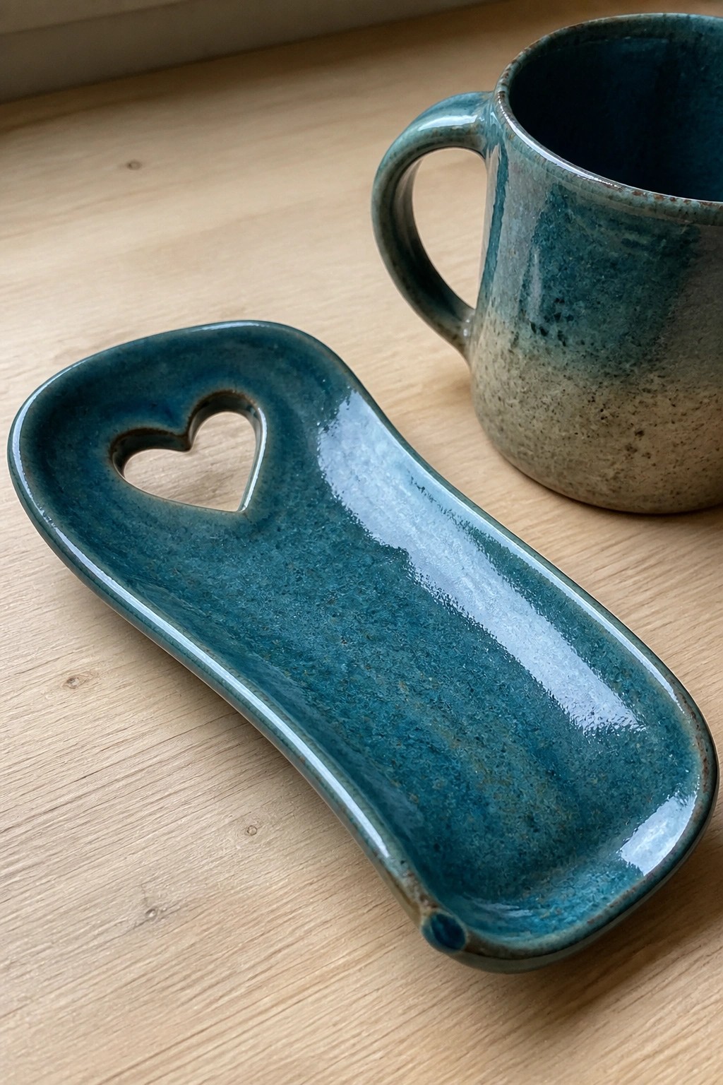 Teal ceramic spoon rest with heart cutout beside matching speckled mug on wood