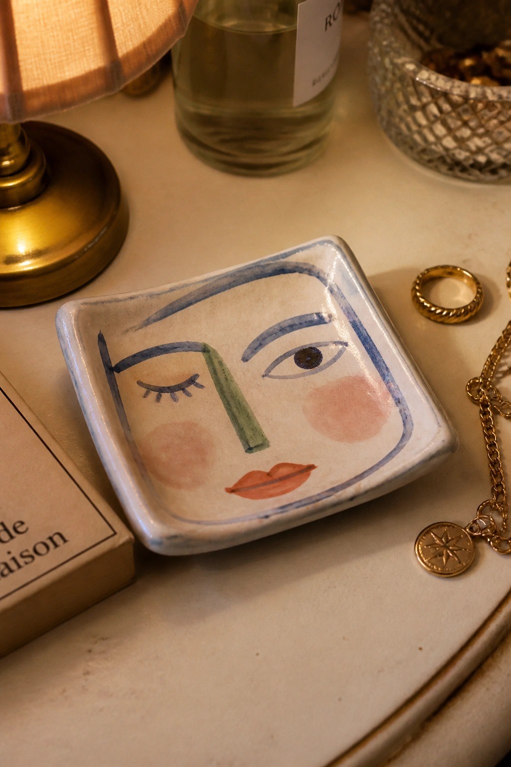 Ceramic tray with abstract face design amid gold jewelry on a table