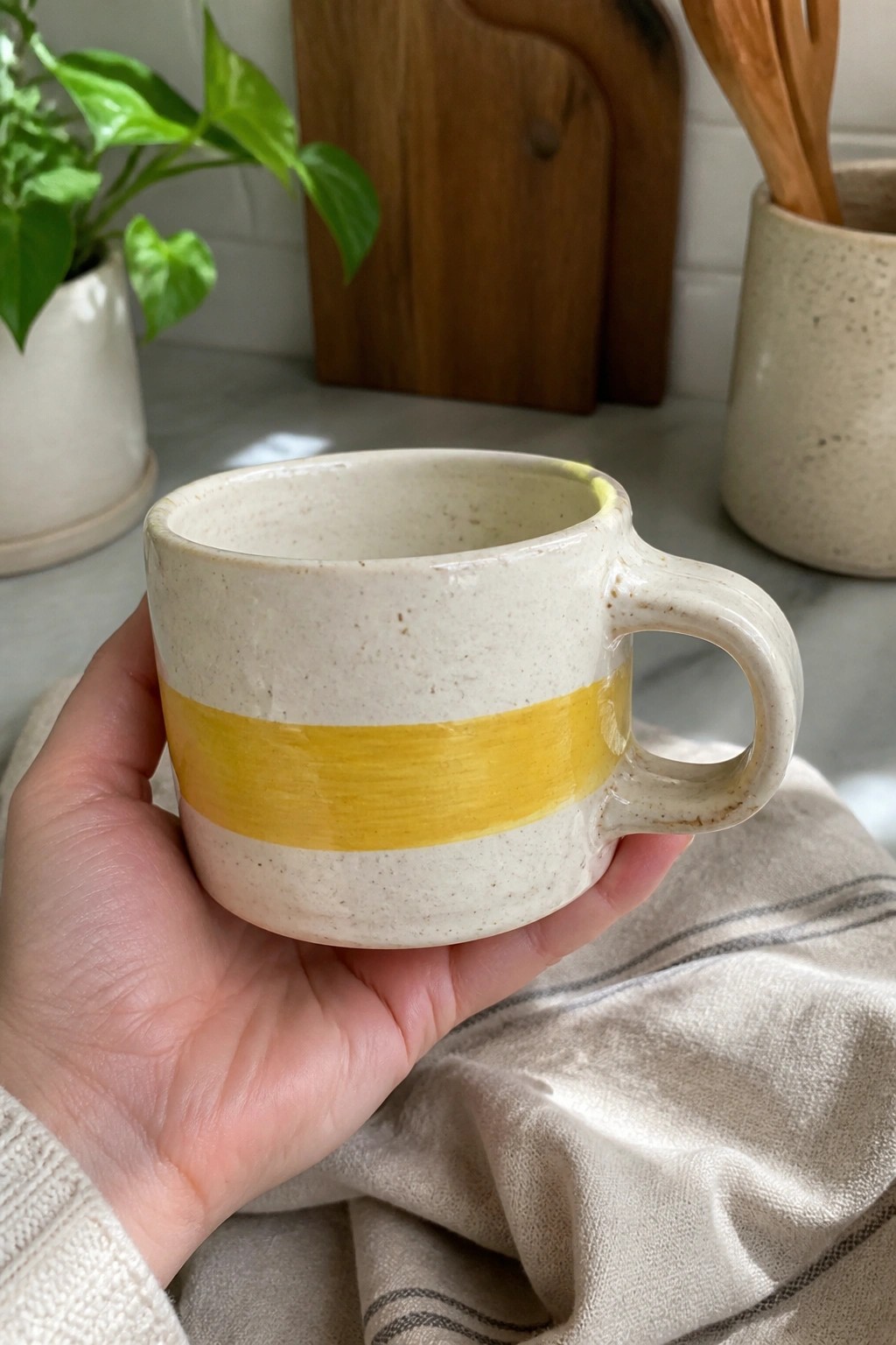 Hand holding speckled off-white mug with yellow stripe over linen cloth
