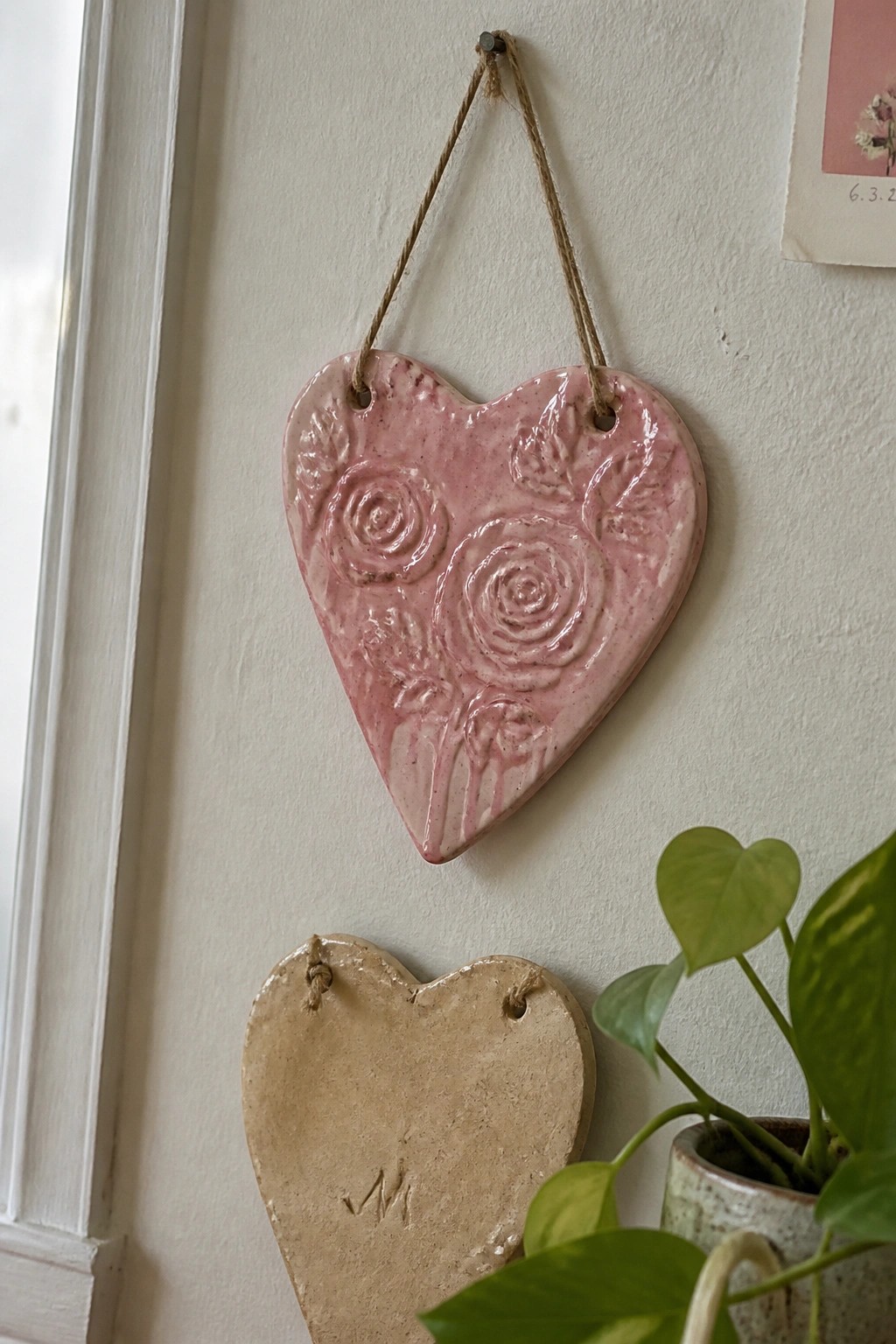 Pink rose-embossed ceramic heart hanging above beige heart on white wall with plant
