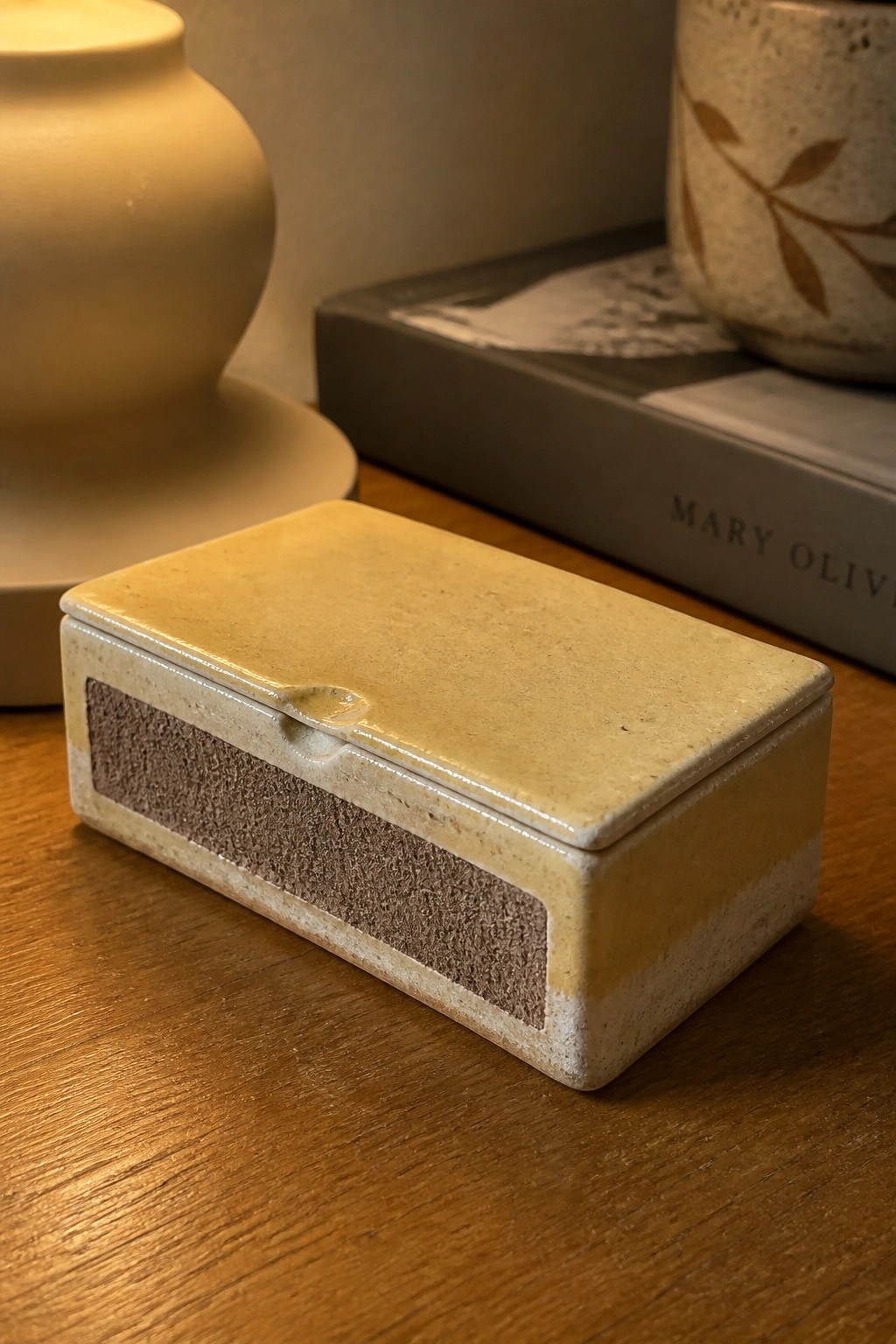 Beige ceramic lidded box with brown band on wooden table near books and vases