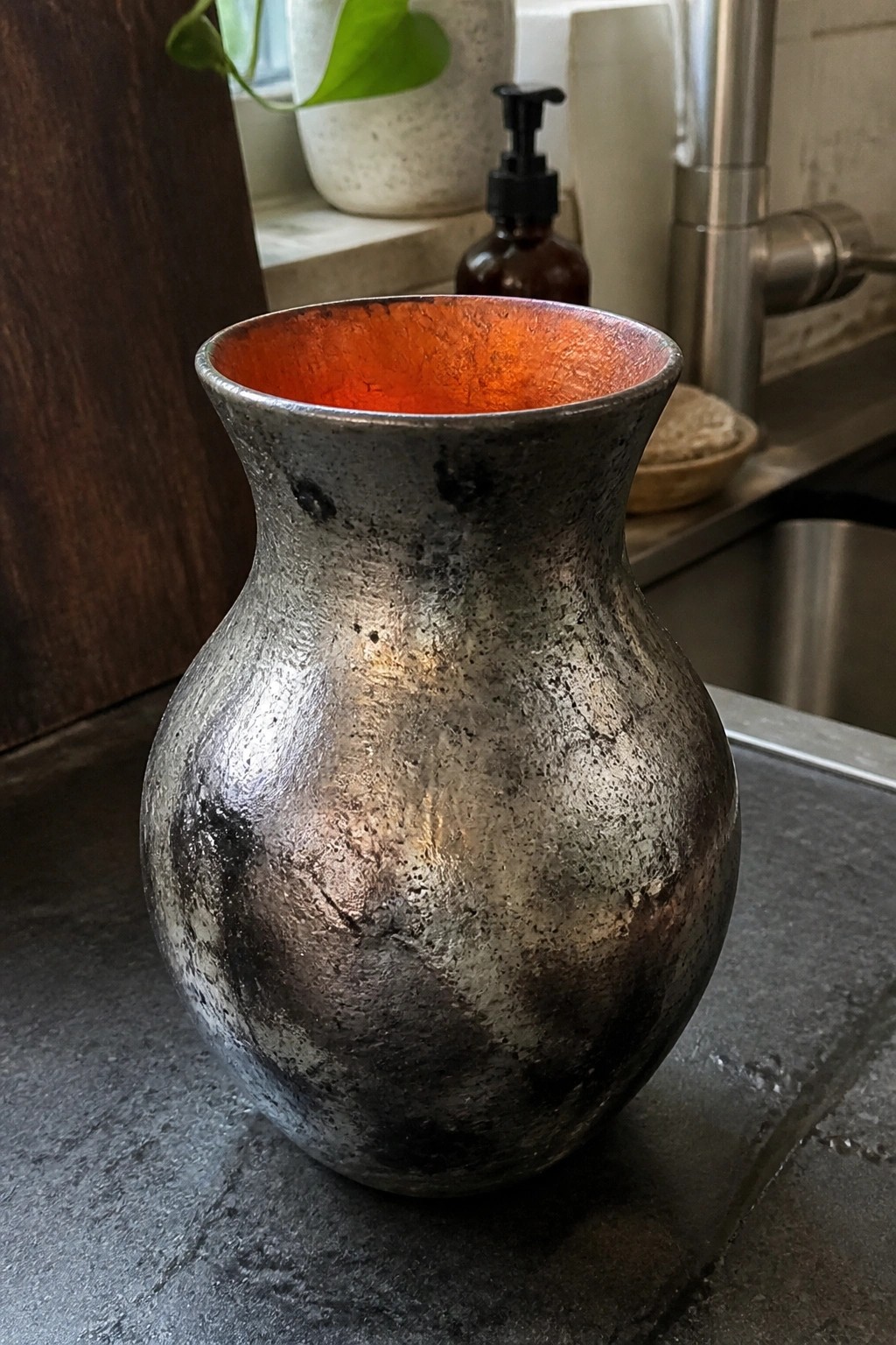 Bulbous metal vase with textured gray exterior and vibrant orange interior on countertop