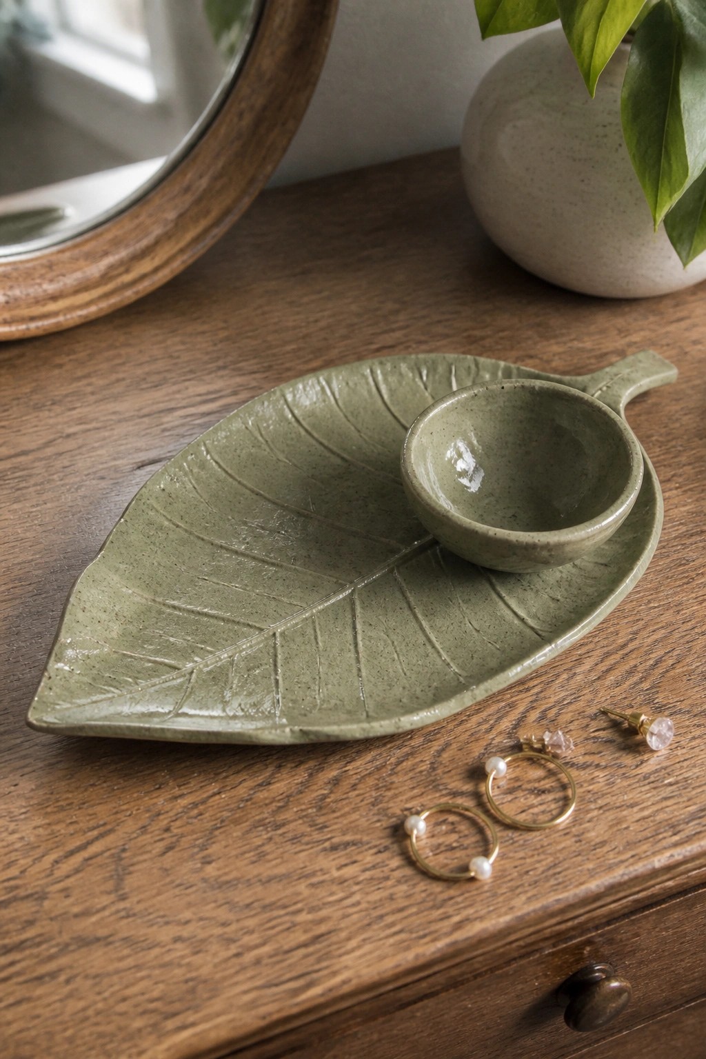 Green ceramic leaf tray with bowl and gold pearl earrings on wooden table.