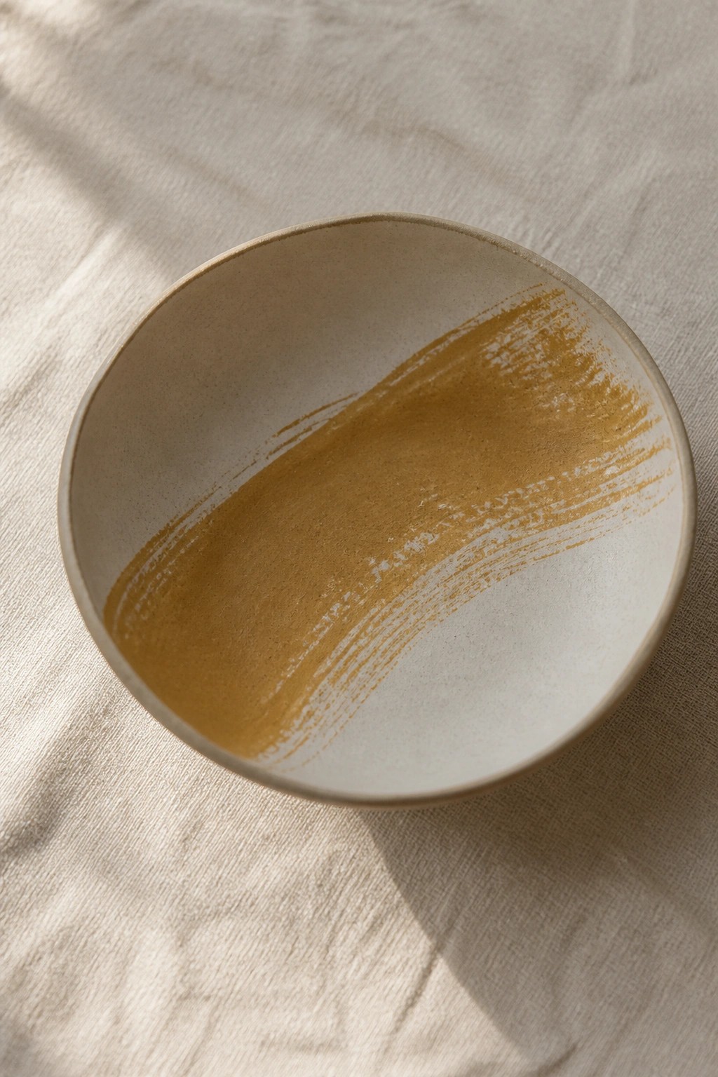 Beige ceramic bowl with sweeping ochre brushstroke on textured linen cloth
