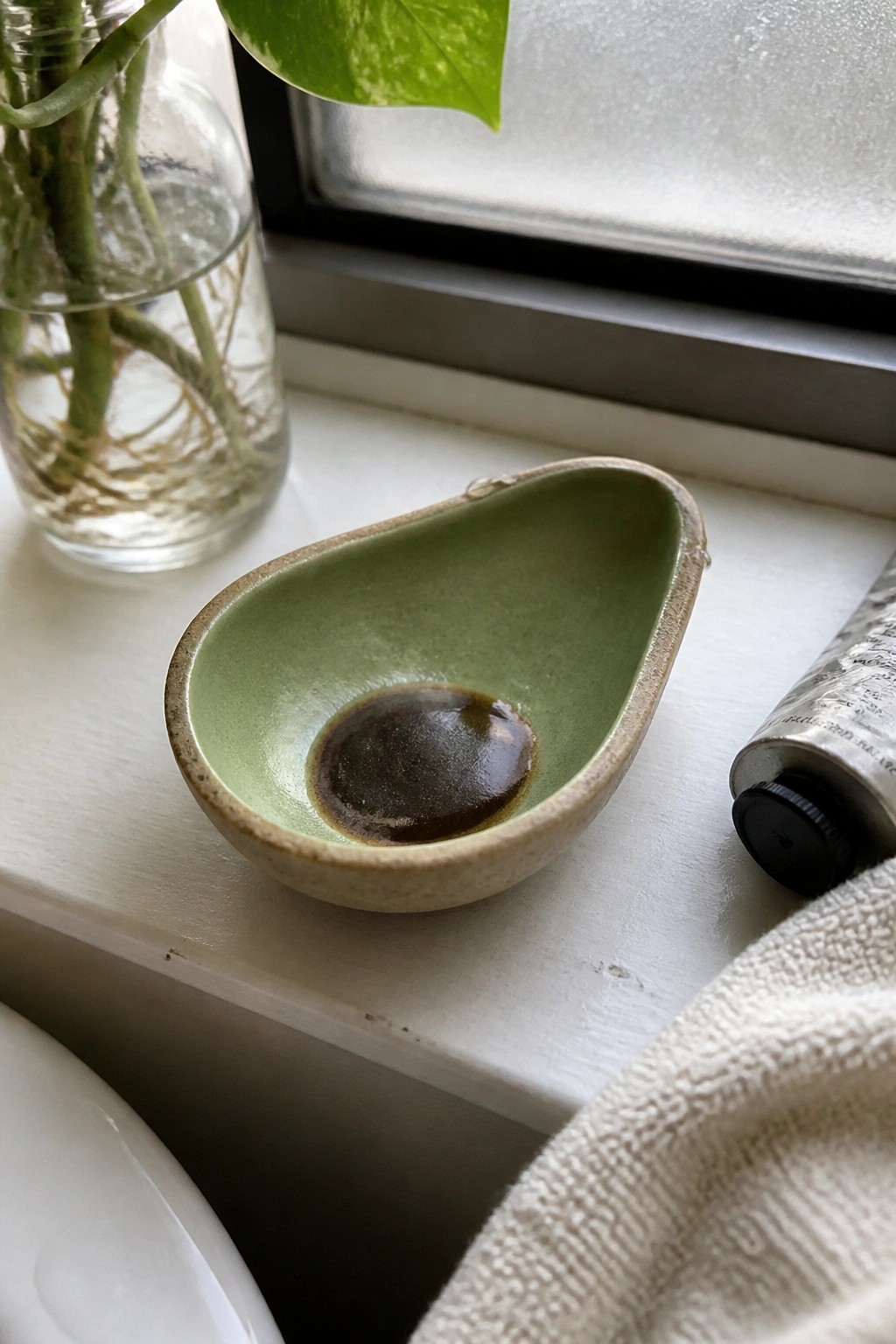 Green ceramic bowl with dark liquid on white windowsill near plant vase and tube