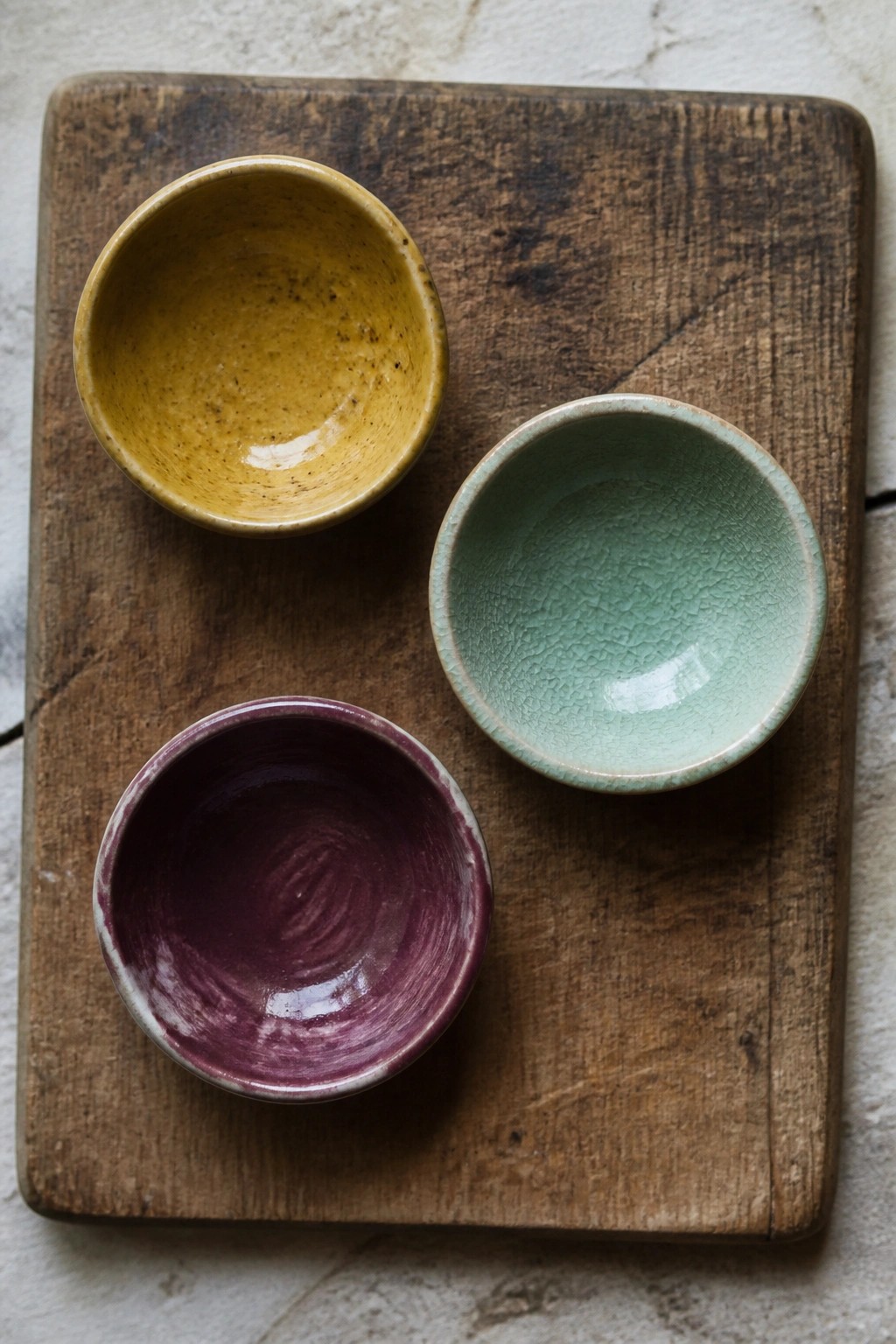 Three colorful ceramic bowls in yellow, green, and purple on a wooden board.