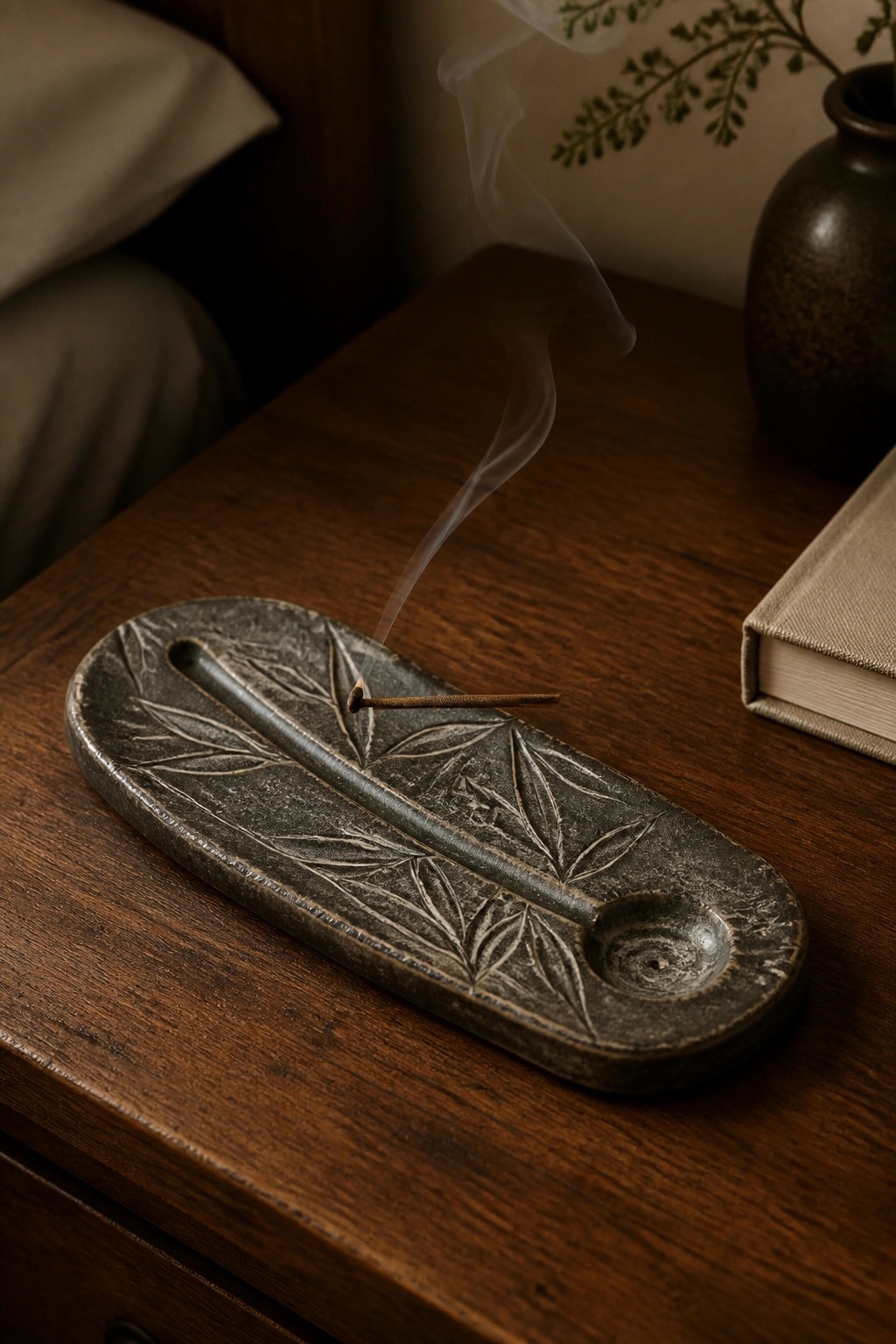 Burning incense stick on leaf-embossed dark ceramic holder atop wooden table with rising smoke.