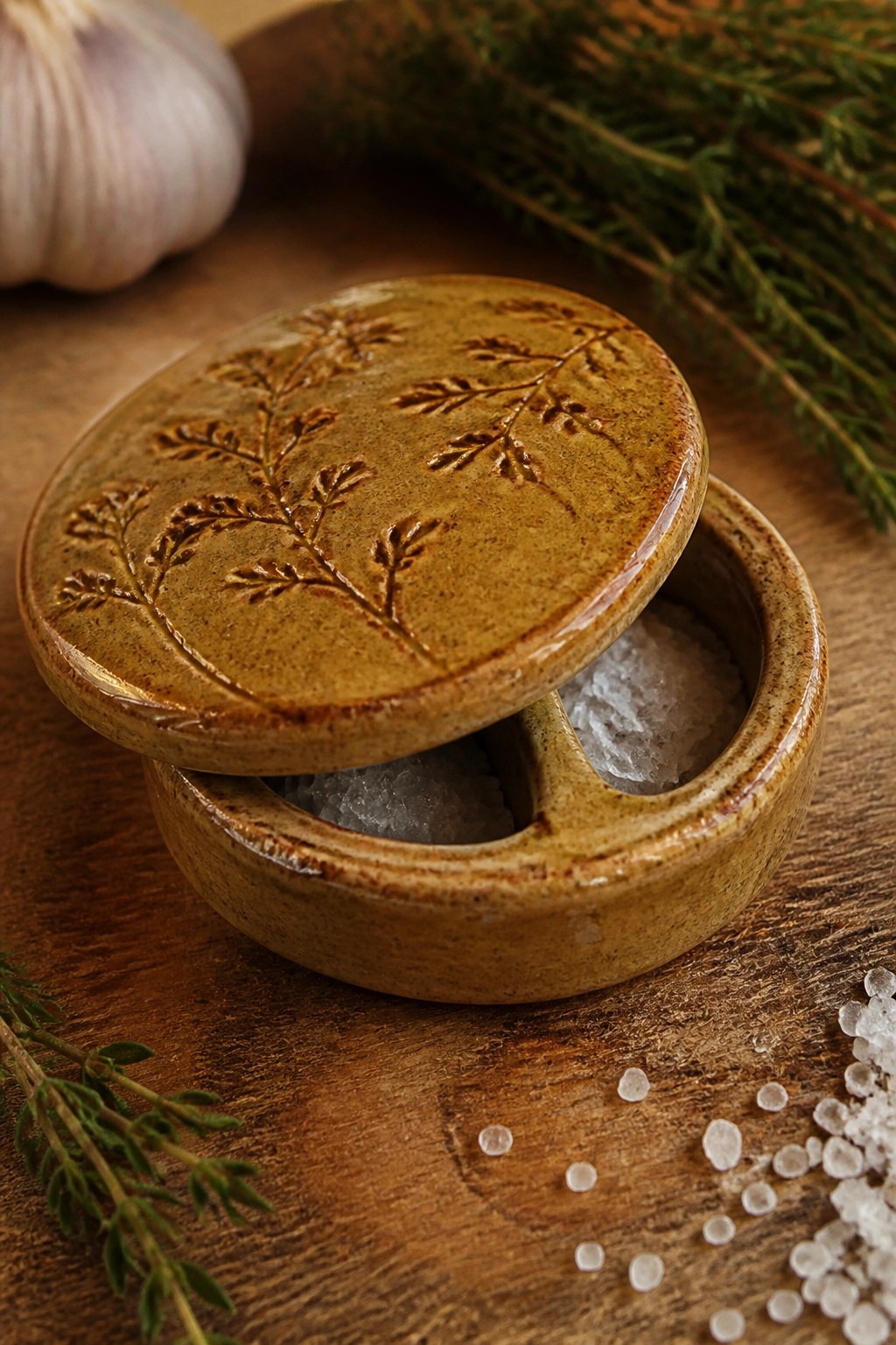 A round ceramic salt cellar with a lidded top decorated by pressed leaf textures sits open on a wooden surface next to scattered salt and fresh herbs.
