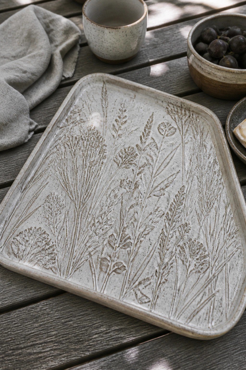 Ceramic tray with embossed wildflowers on wooden table beside gray linen and bowls