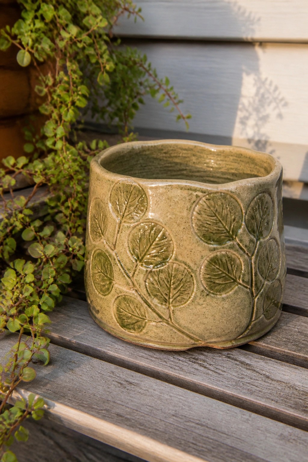 Green glazed ceramic bowl with embossed leaf designs on wooden slats among plants.