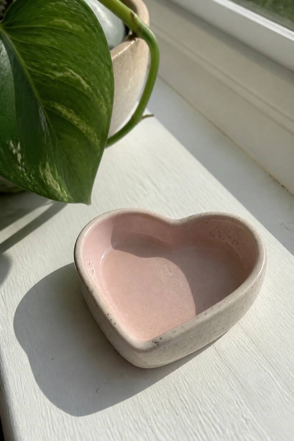 Pink heart-shaped ceramic bowl on sunlit white windowsill with green plant leaf