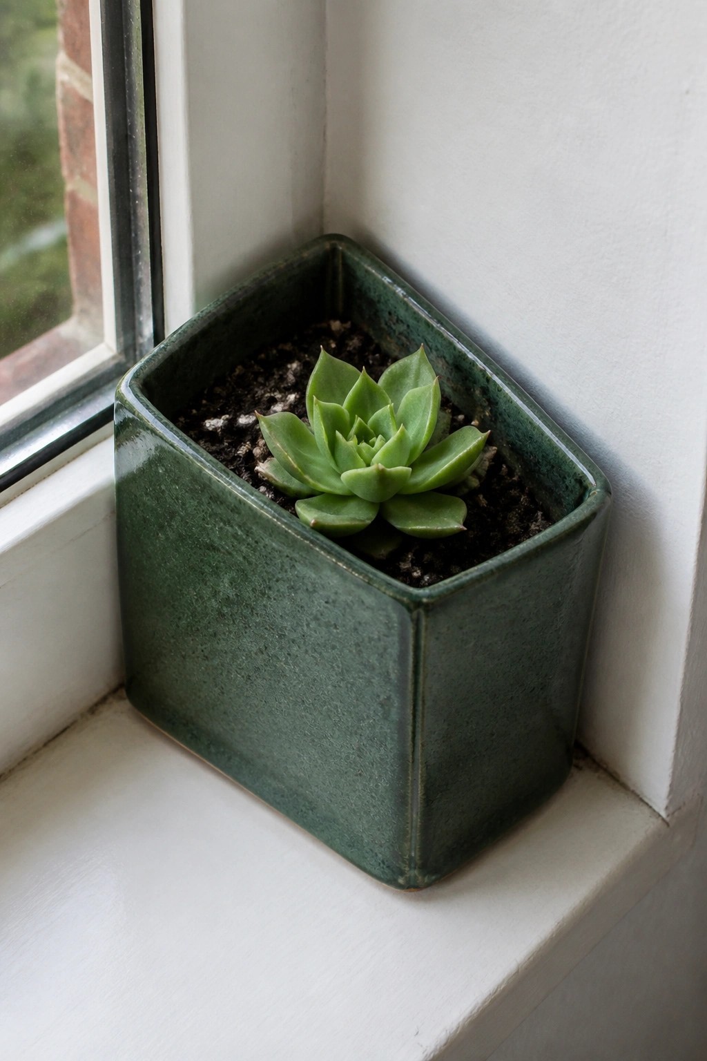 A small green ceramic planter holding a succulent sits on a white windowsill next to a window.