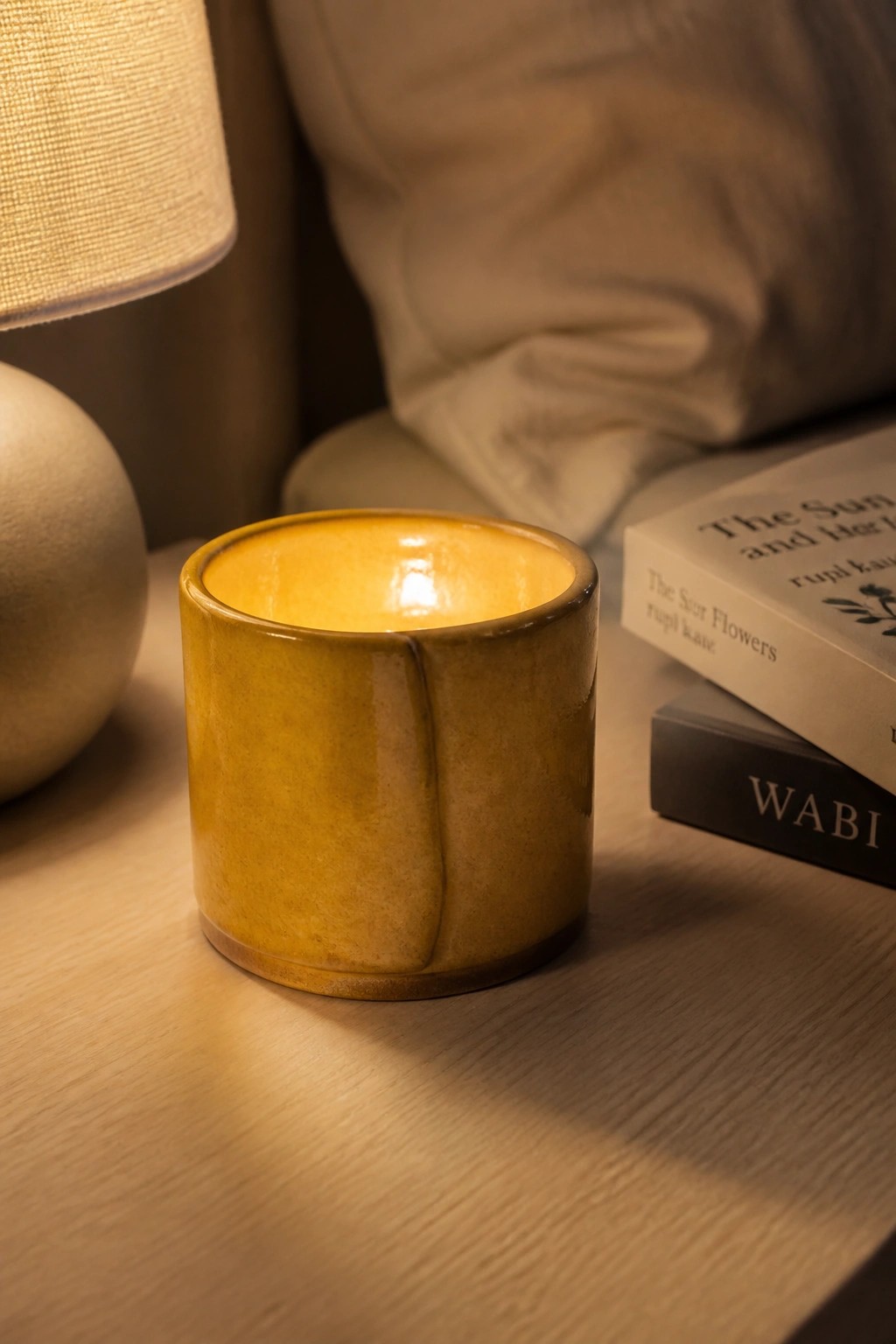 Yellow ceramic candle glowing on wooden table beside books and lamp
