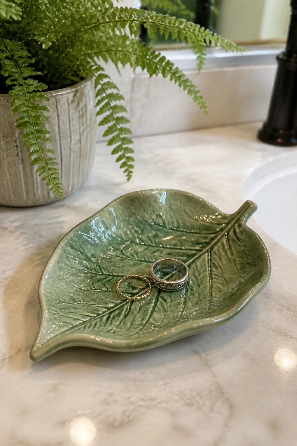 Green ceramic leaf dish holding two rings on a white marble counter next to a potted fern.