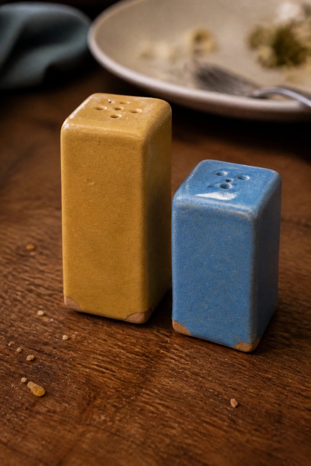 Two rectangular ceramic salt and pepper shakers, one yellow and one blue, rest on a wooden table beside a plate.