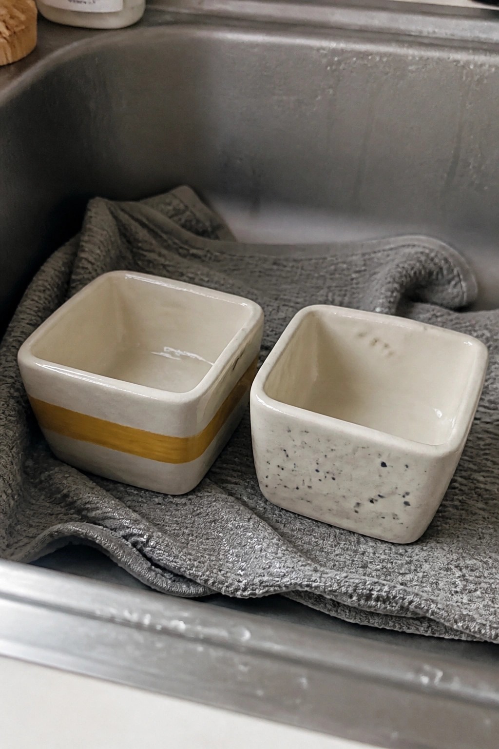 Two square ceramic bowls, one yellow-striped and one speckled, on gray towel in sink.