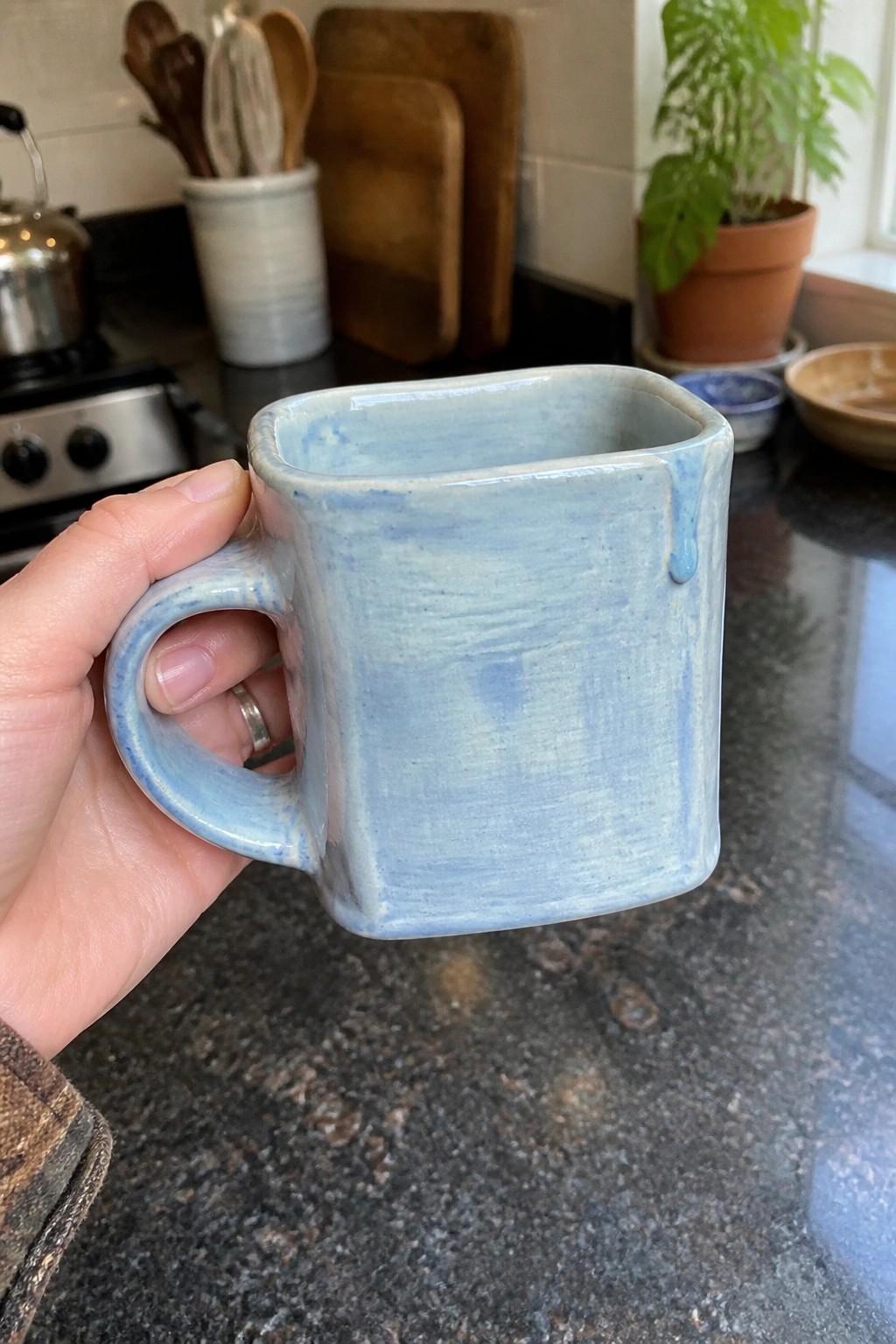 Hand in plaid sleeve holding light blue square ceramic mug in kitchen.