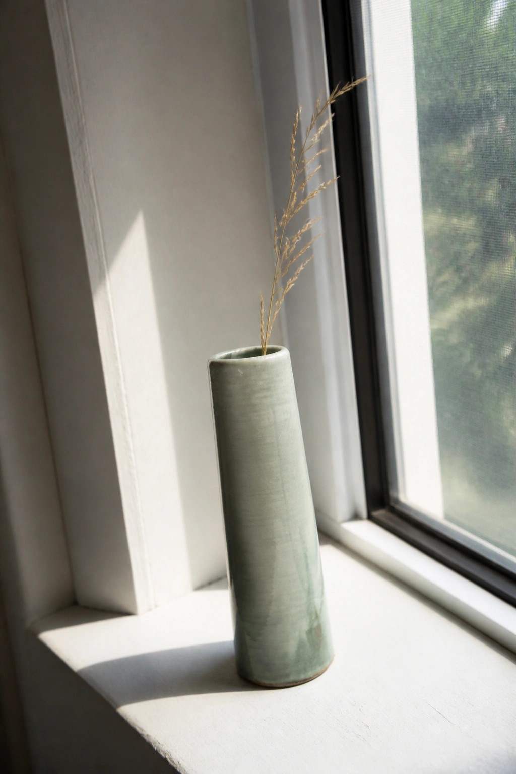Green ceramic vase with dried grass on sunlit white windowsill.