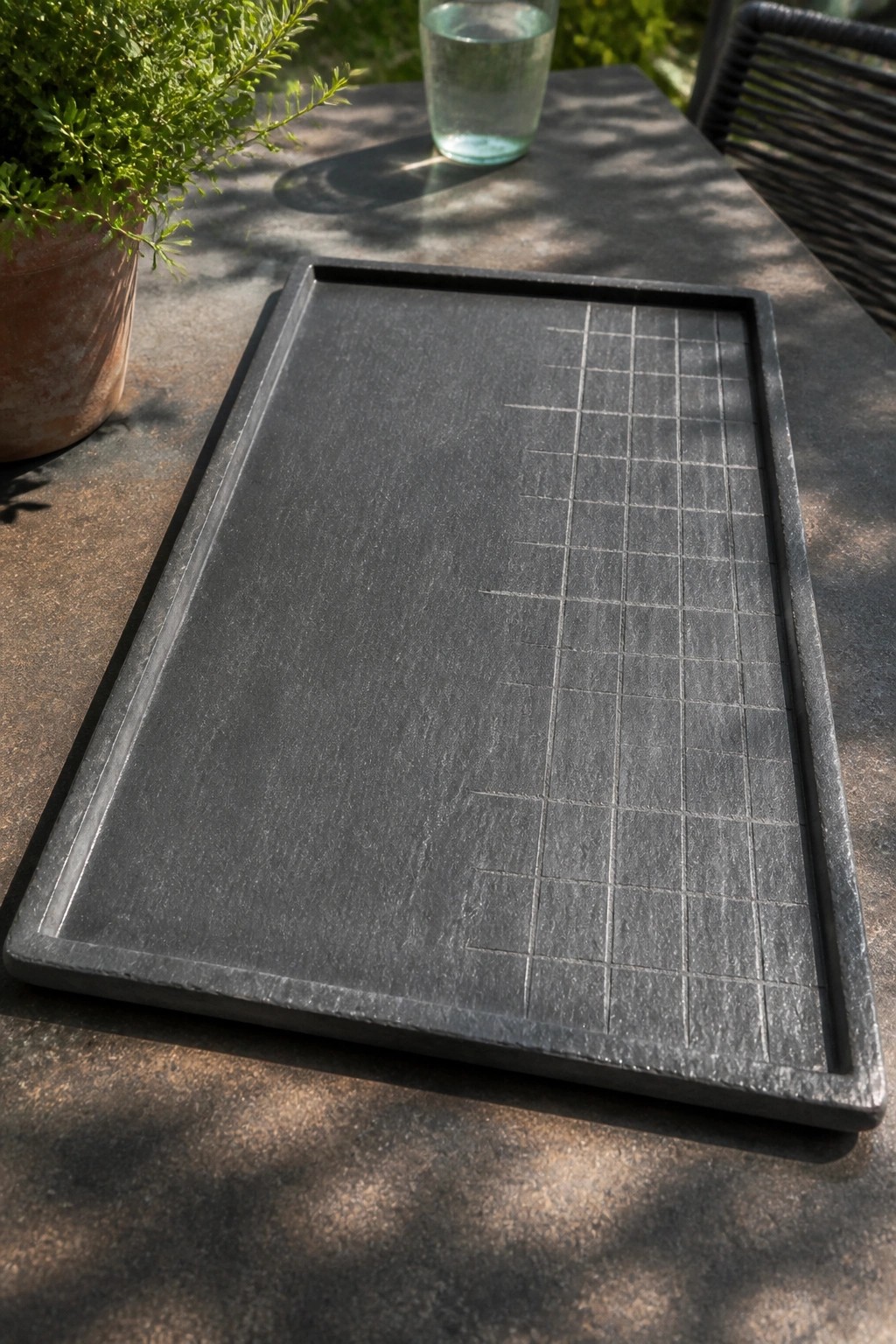 Black rectangular tray with grid on stone table beside glass and potted plant