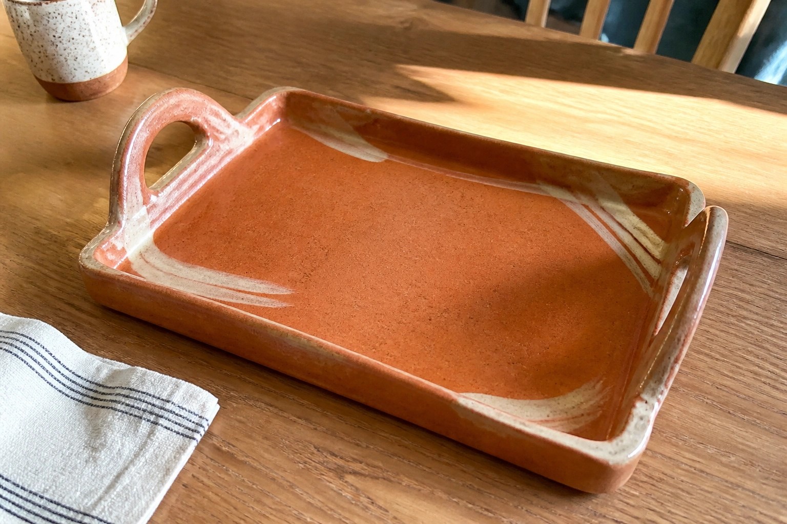Orange ceramic tray with handles on sunlit wooden table beside beige mug