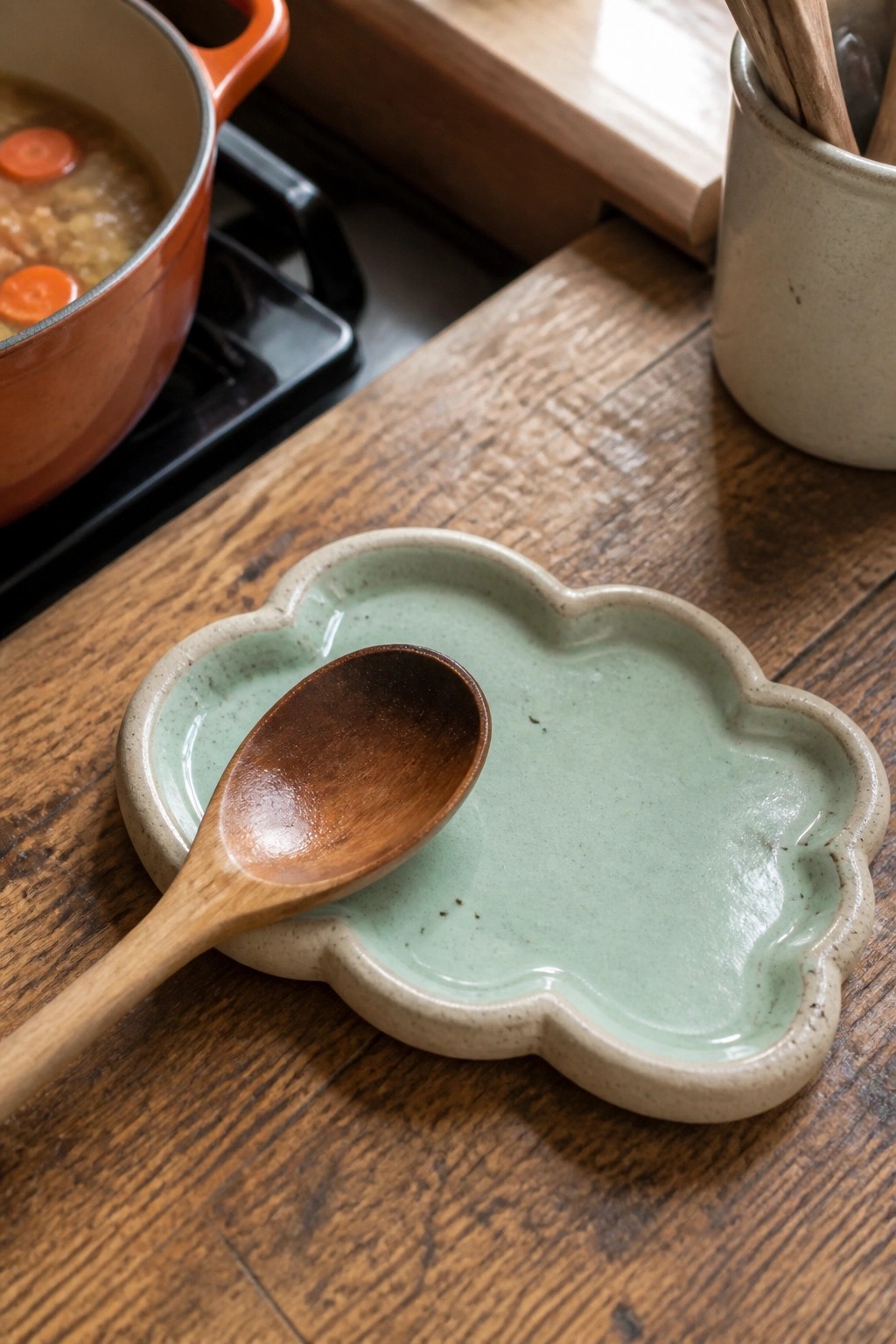 Wooden spoon on light green scalloped ceramic dish beside orange pot on stove.