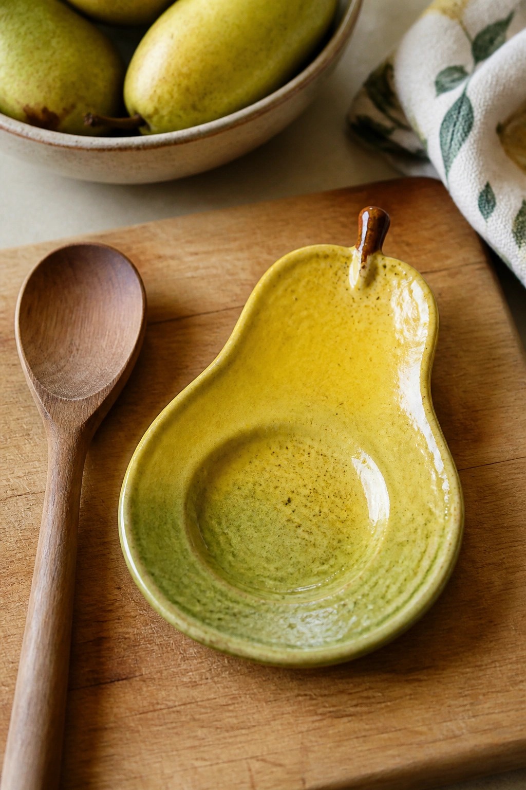 Yellow-green pear-shaped ceramic dish on wooden board beside spoon and pears