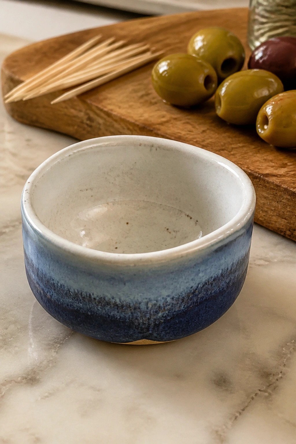Blue ceramic bowl on marble beside olives and toothpicks on wooden board