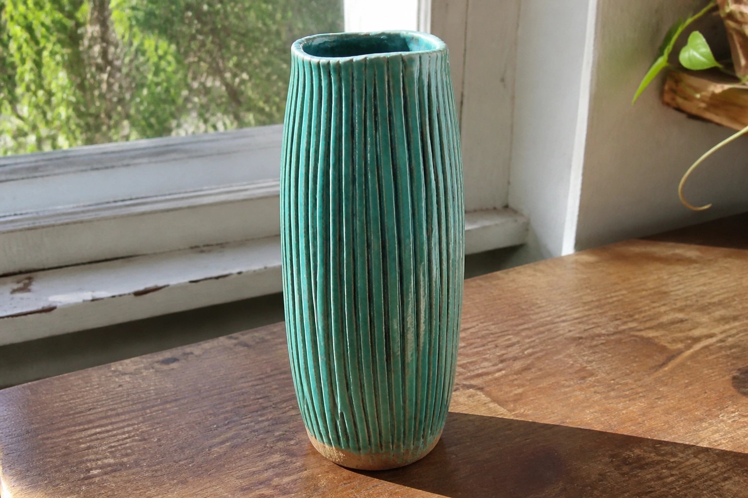 Tall teal ribbed ceramic vase on wooden windowsill with potted plants