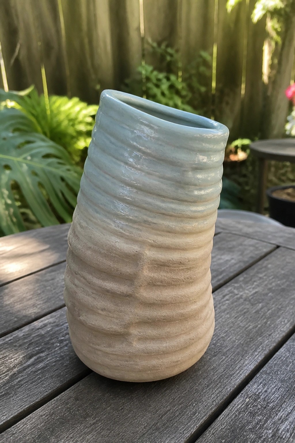 Light blue and beige ridged ceramic vase on wooden outdoor table with plants behind.