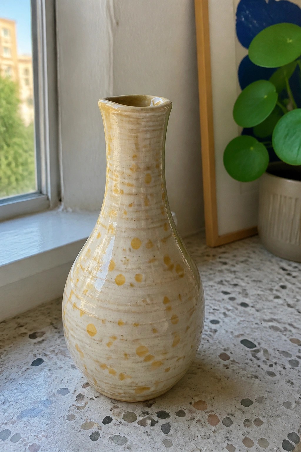 A handmade ceramic vase with a bulbous body, narrow neck, cream and yellow speckled glaze, and horizontal texture lines sits on a terrazzo surface near a window.