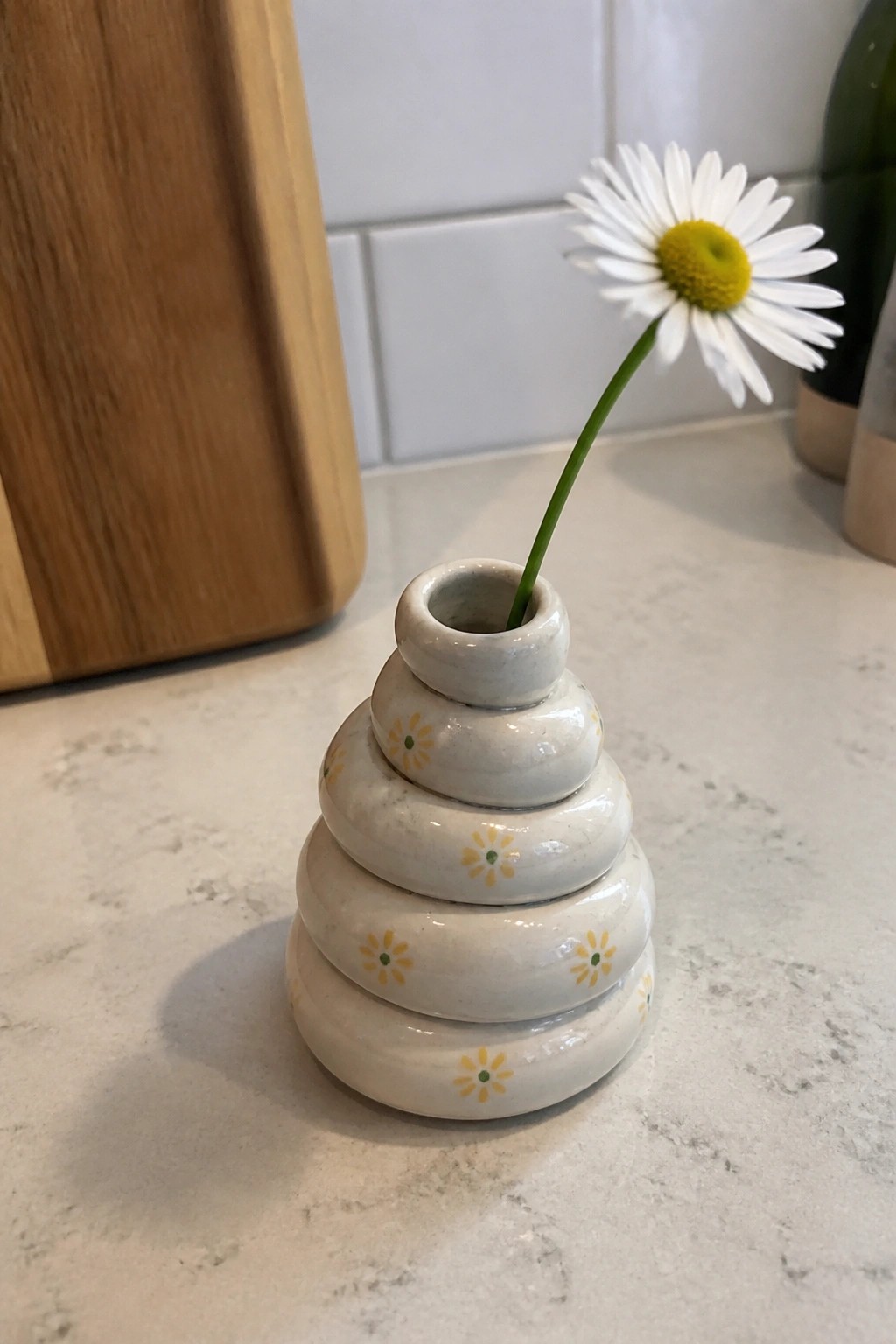 Tiered white ceramic vase with yellow daisies holding one fresh daisy on marble counter.