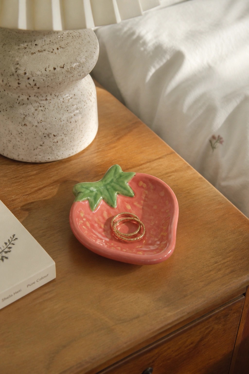 Pink strawberry ceramic dish holding gold rings on wooden nightstand beside vase and book