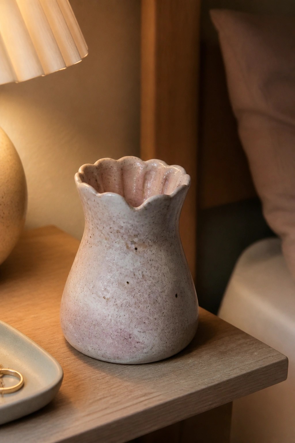 Pink speckled ceramic vase with scalloped rim on wooden nightstand beside lamp