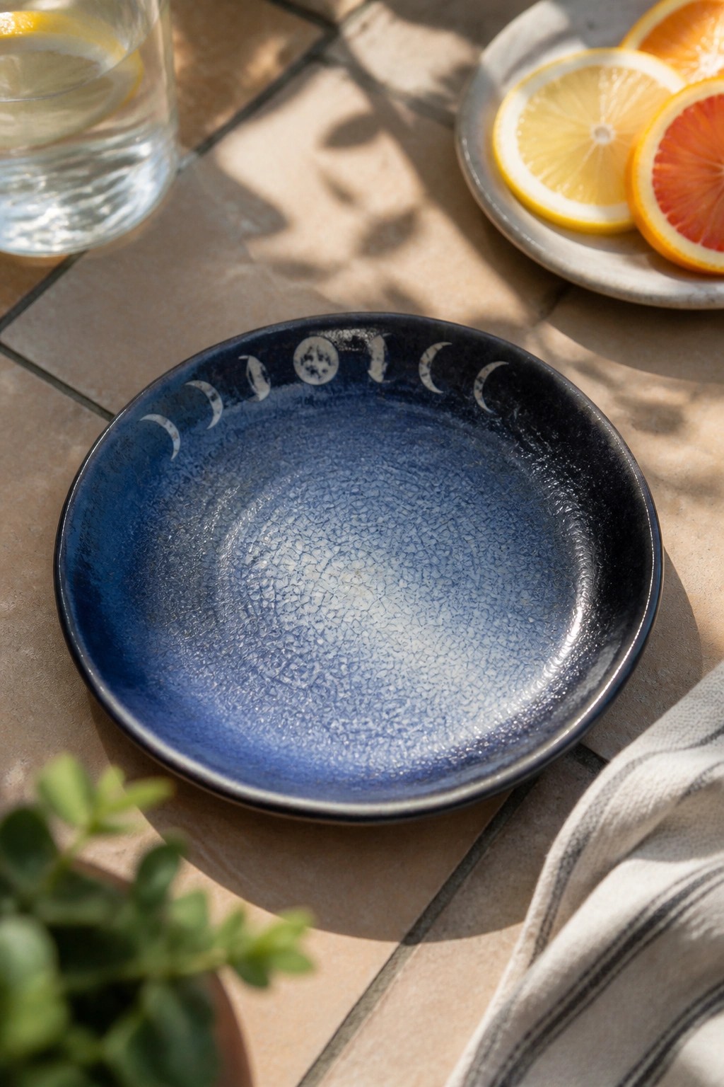 Blue crackled plate with moon phases beside citrus slices and lemon water on tiles.