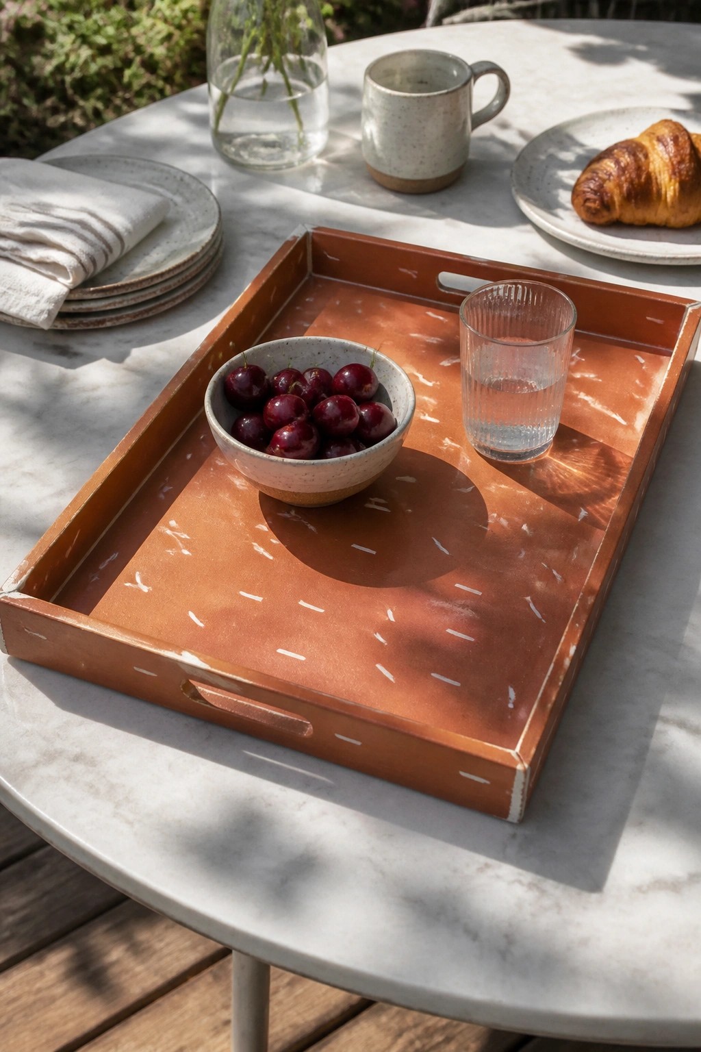 Terracotta tray with bowl of cherries and water glass beside croissant on marble table
