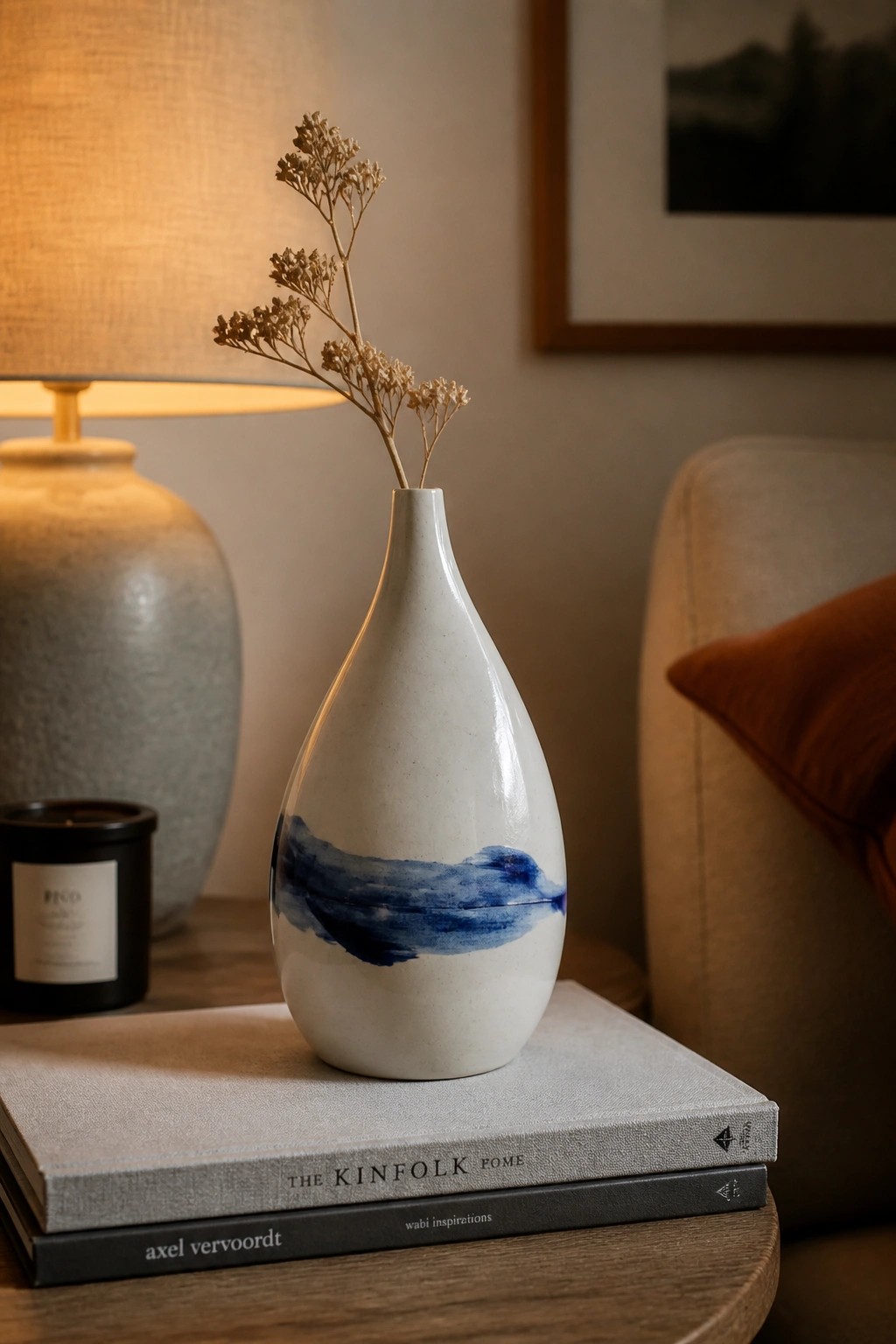 White ceramic vase with blue accents holding dried flowers on stacked books beside lit lamp