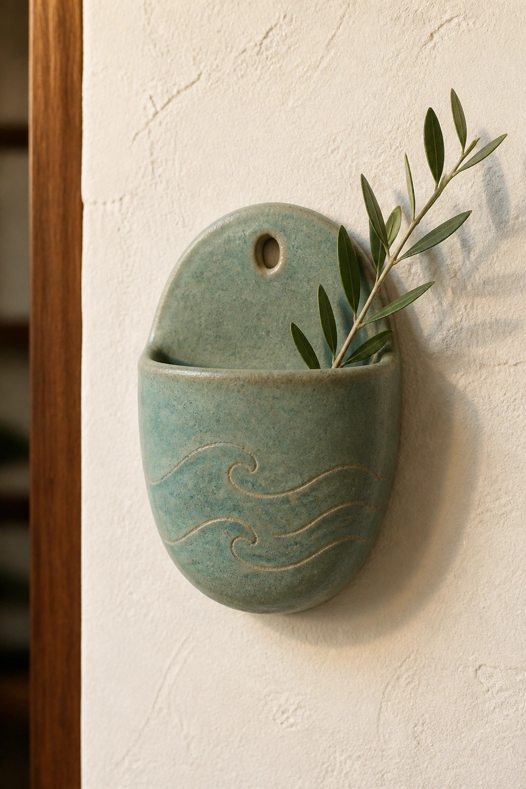Teal ceramic wall pocket with wave patterns holding olive branch on white wall.
