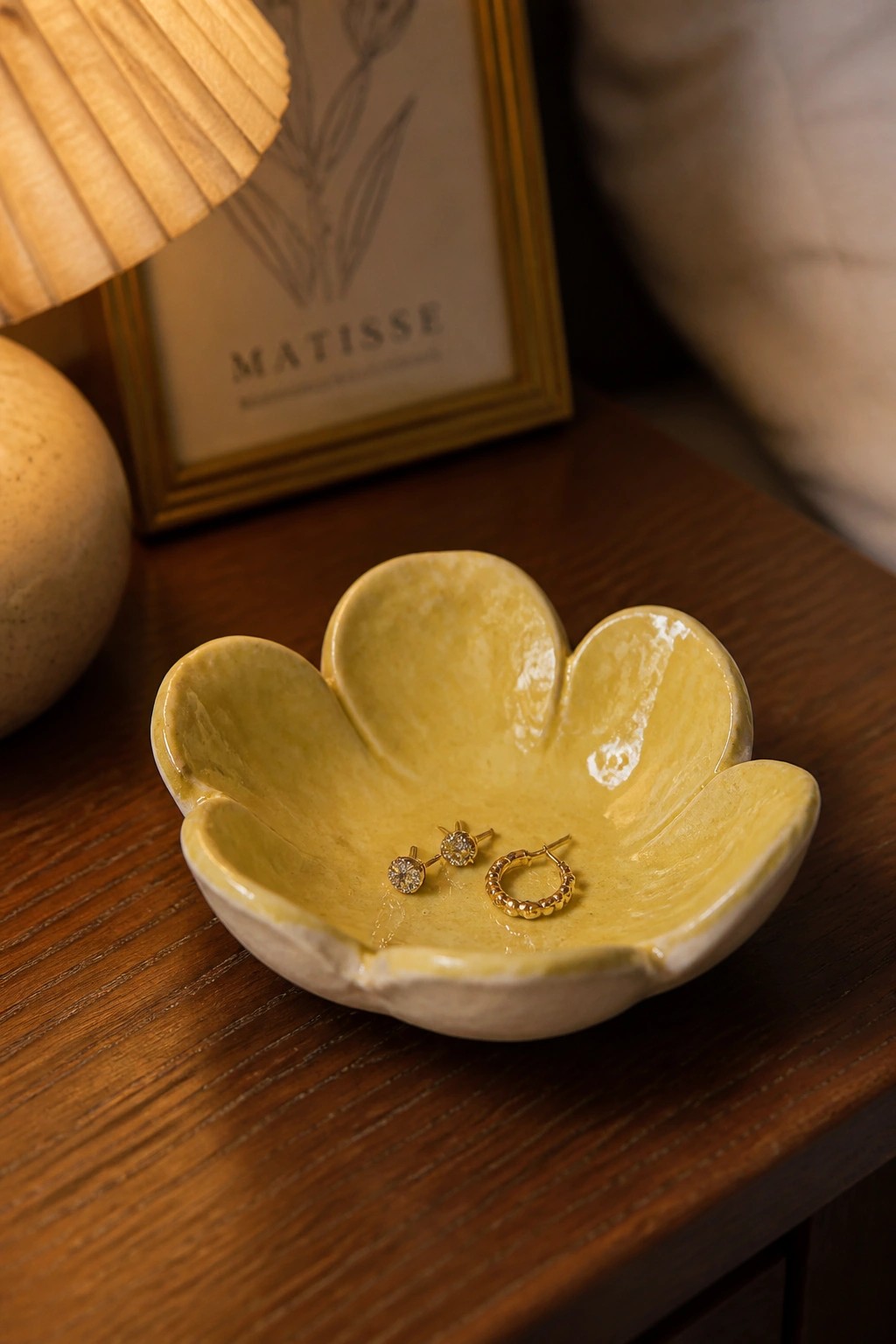 Yellow ceramic flower bowl holding gold earrings on wooden table with Matisse print.