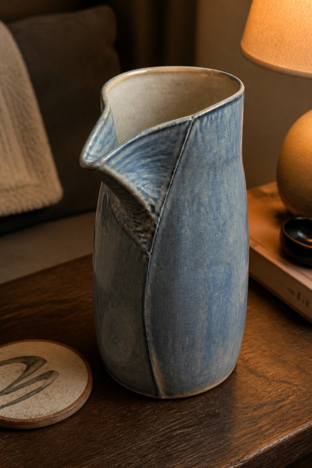 Blue ceramic pitcher with spout on wooden table beside lamp and book