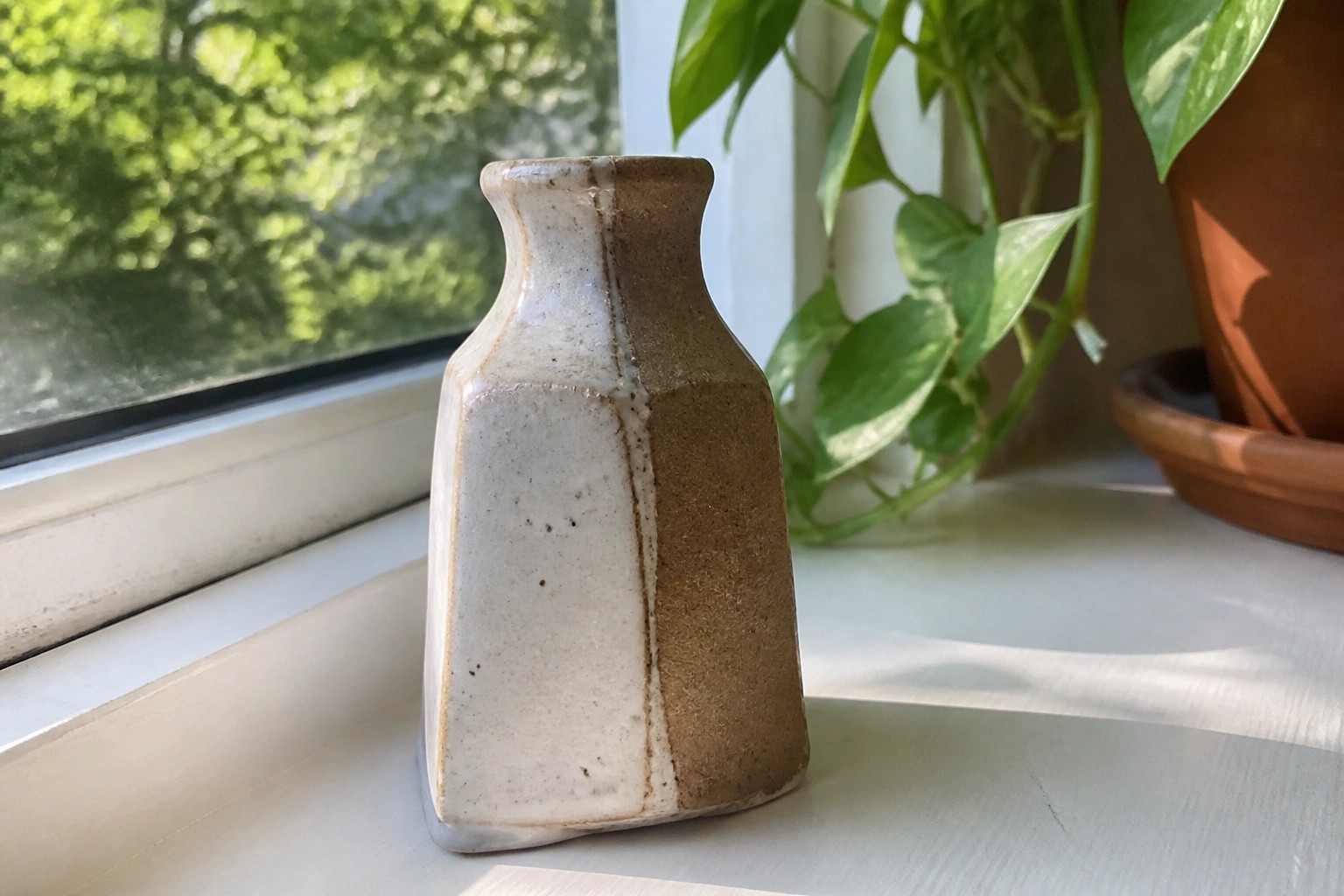 Speckled white ceramic vase with brown unglazed side on sunny windowsill with plants.