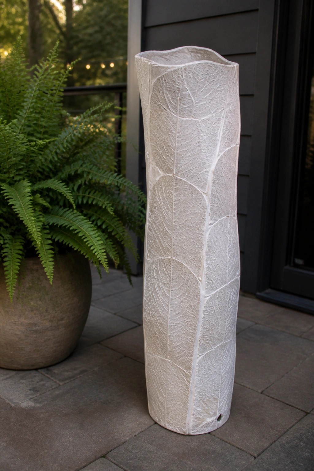 Tall white leaf-textured vase beside potted green fern on stone patio