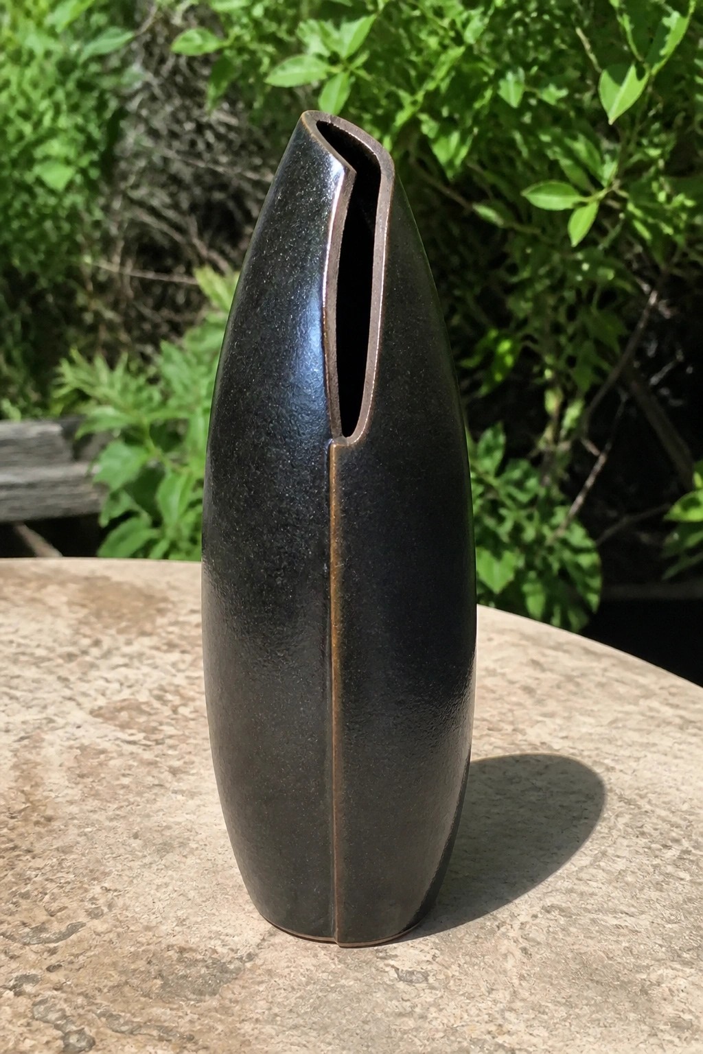 Tall dark bronze vase with vertical slit on stone table outdoors